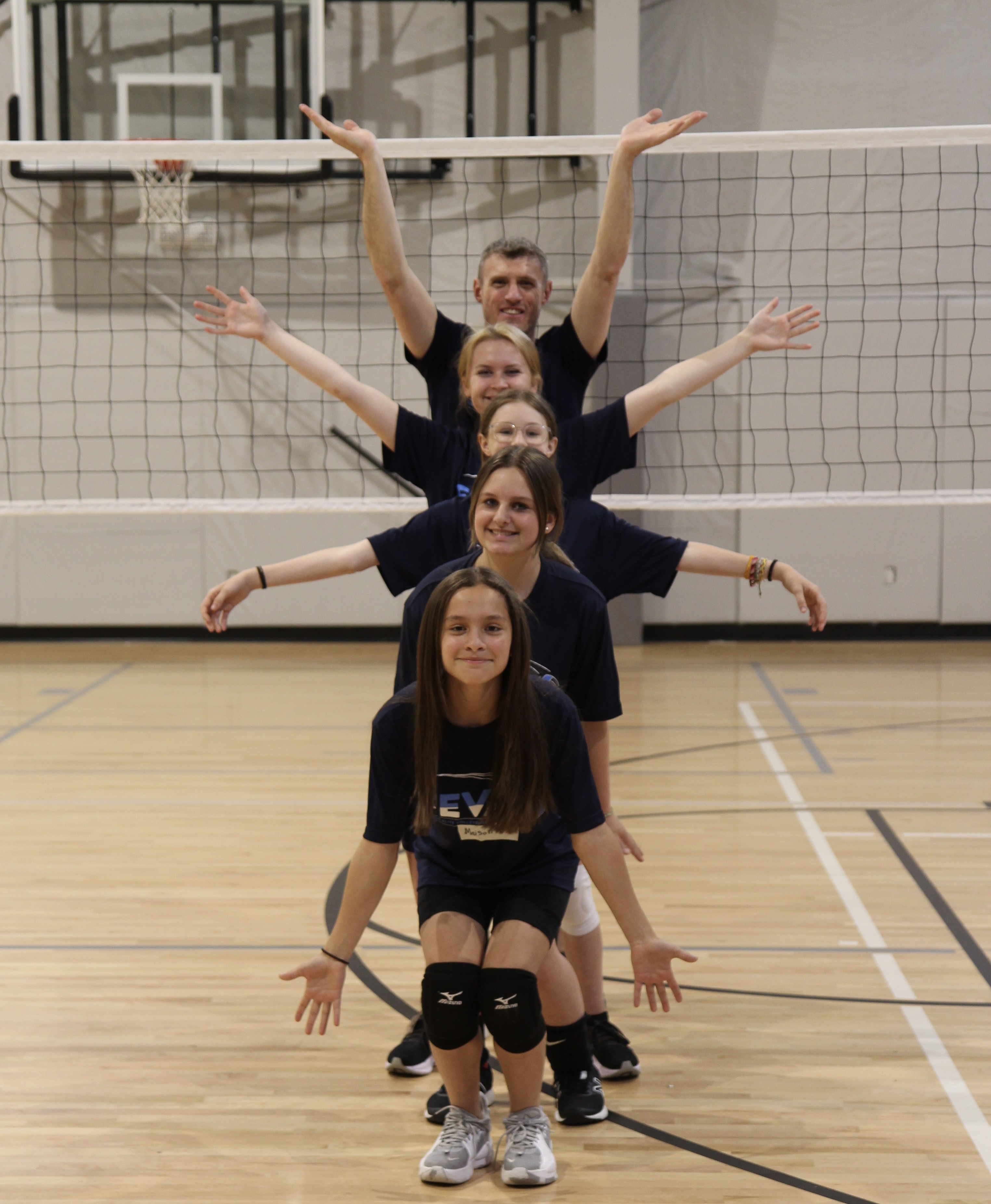 A group of five volleyball players, including a coach, standing in a formation on a gym court, with arms raised in celebration or greeting.