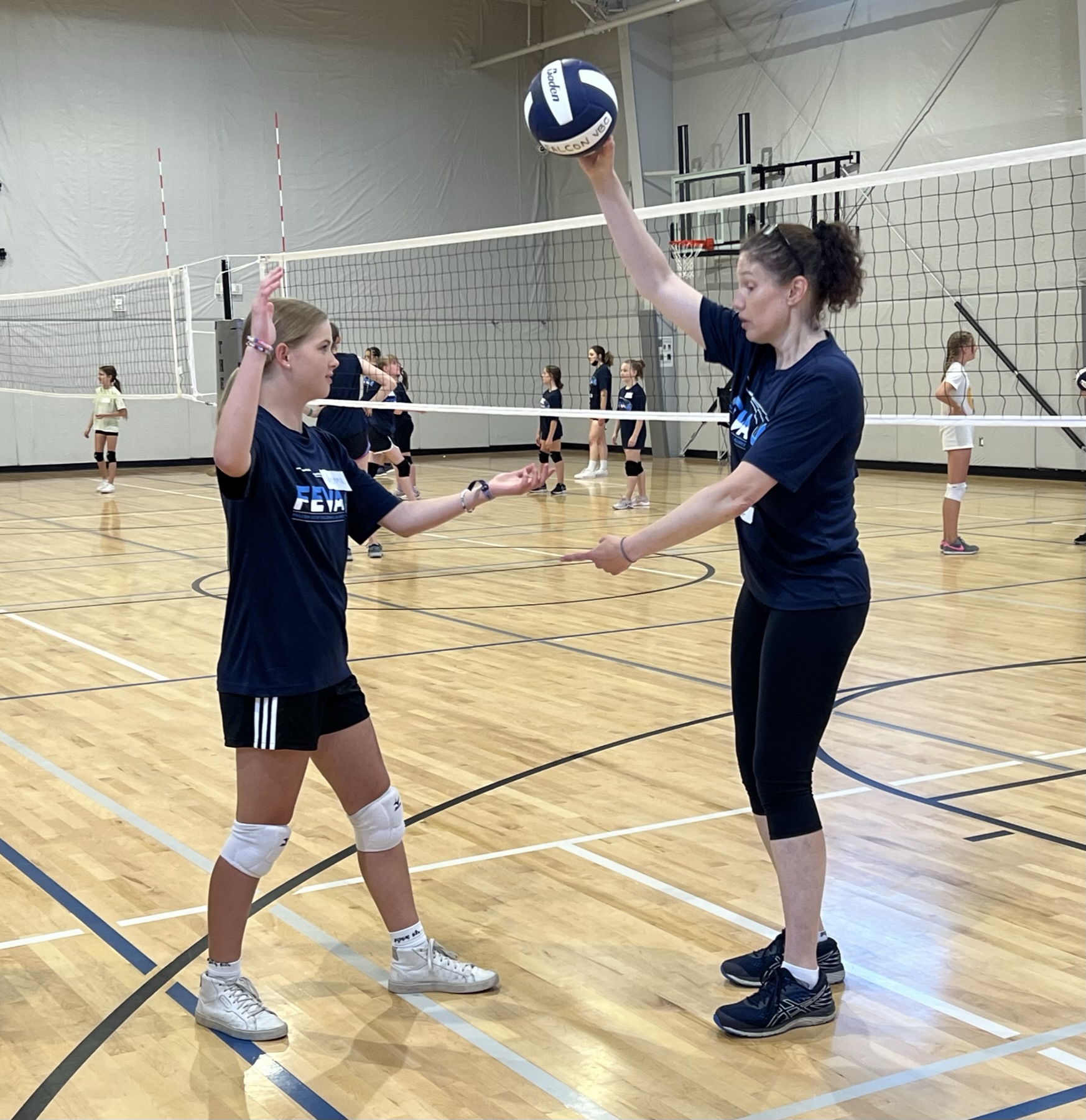 Coach Hammer demonstrates a skill to a student in a gym setting, with a volleyball net in the background and other players practicing.