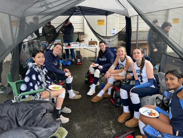 A group of six young female volleyball players from the FEVA 14 team enjoying a meal together under a tent, smiling and relaxing between games at a tournament.