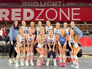 The FEVA volleyball team poses together smiling in front of a red backdrop that reads 'RED ZONE', showcasing their uniforms and team spirit.