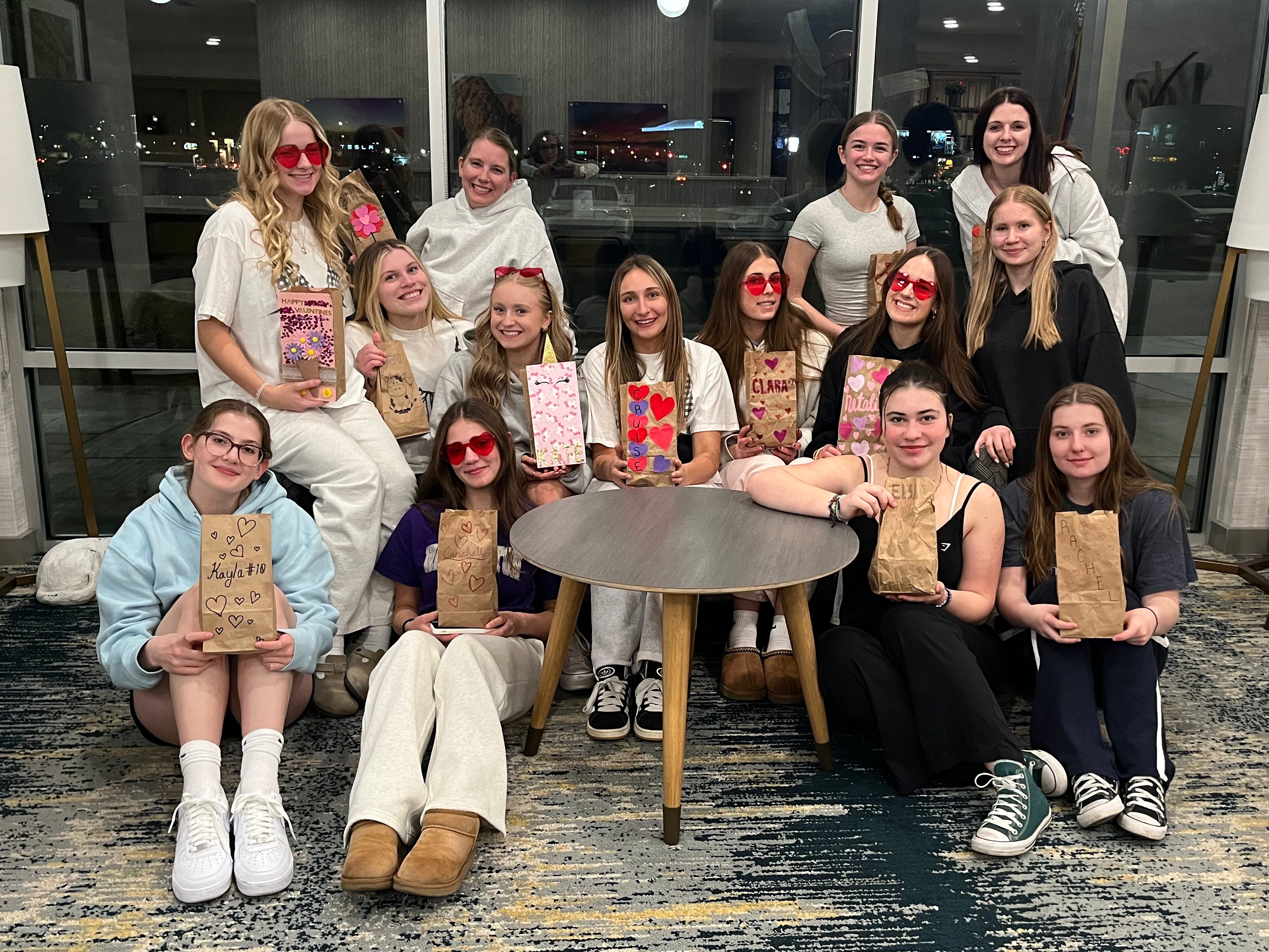 A group of FEVA players in playful attire, wearing heart-shaped sunglasses, sitting around a table. They are holding decorated bags that appear to contain Valentine’s Day gifts, with smiles and a festive atmosphere.