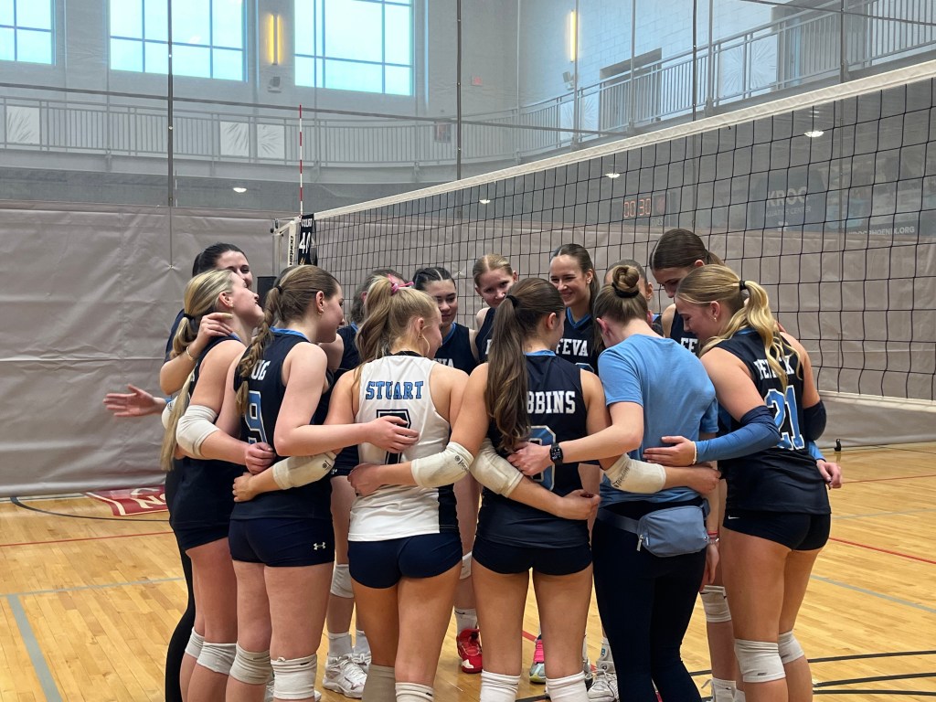 A group of youth FEVA volleyball players huddled together in a gym setting, showing teamwork and camaraderie as they prepare for a match.