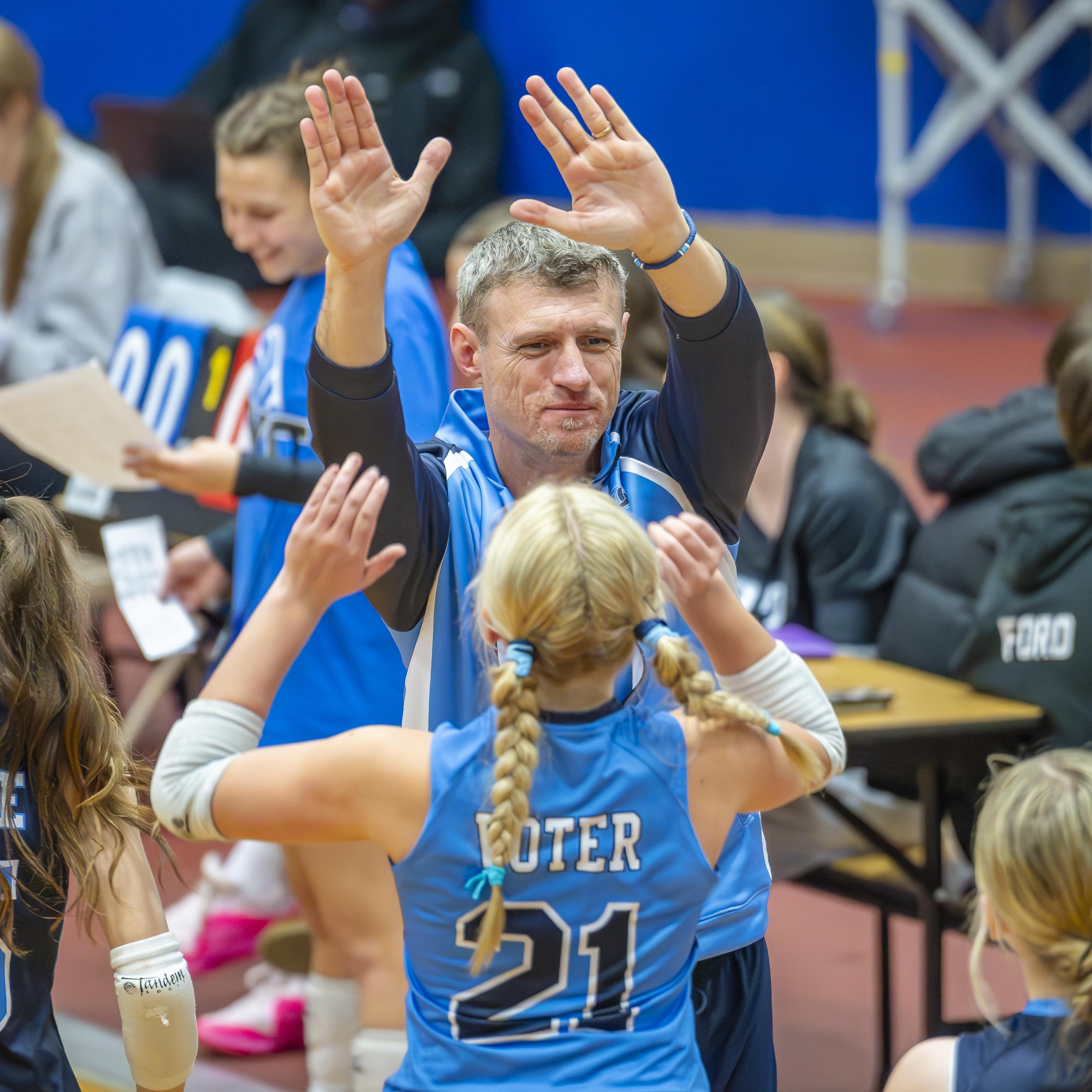 Coach Piper celebrating with players during a volleyball game, showing enthusiasm and support.