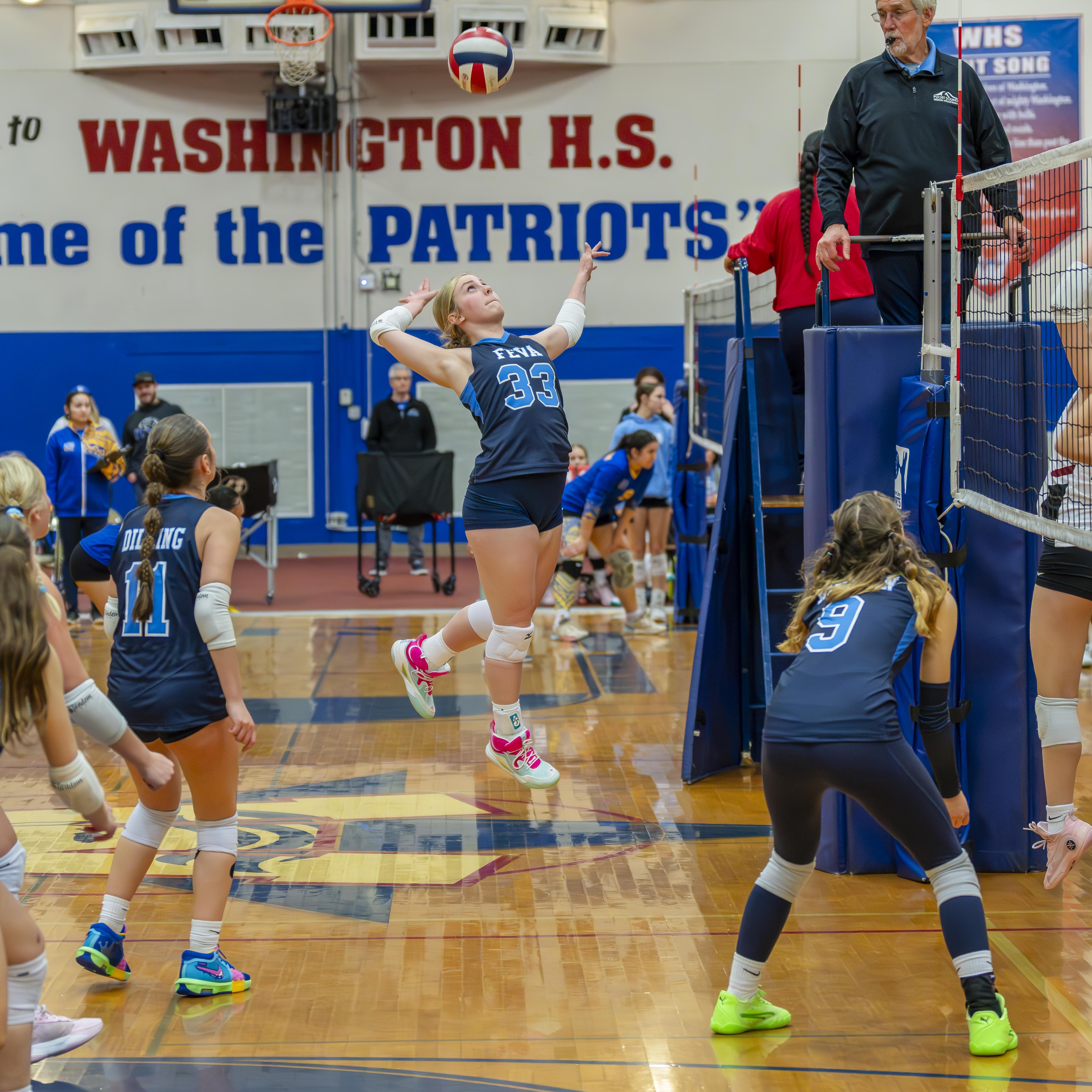 A female volleyball player wearing jersey number 33 prepares to serve the ball during a match at Washington High School, with teammates and a coach visible in the background.