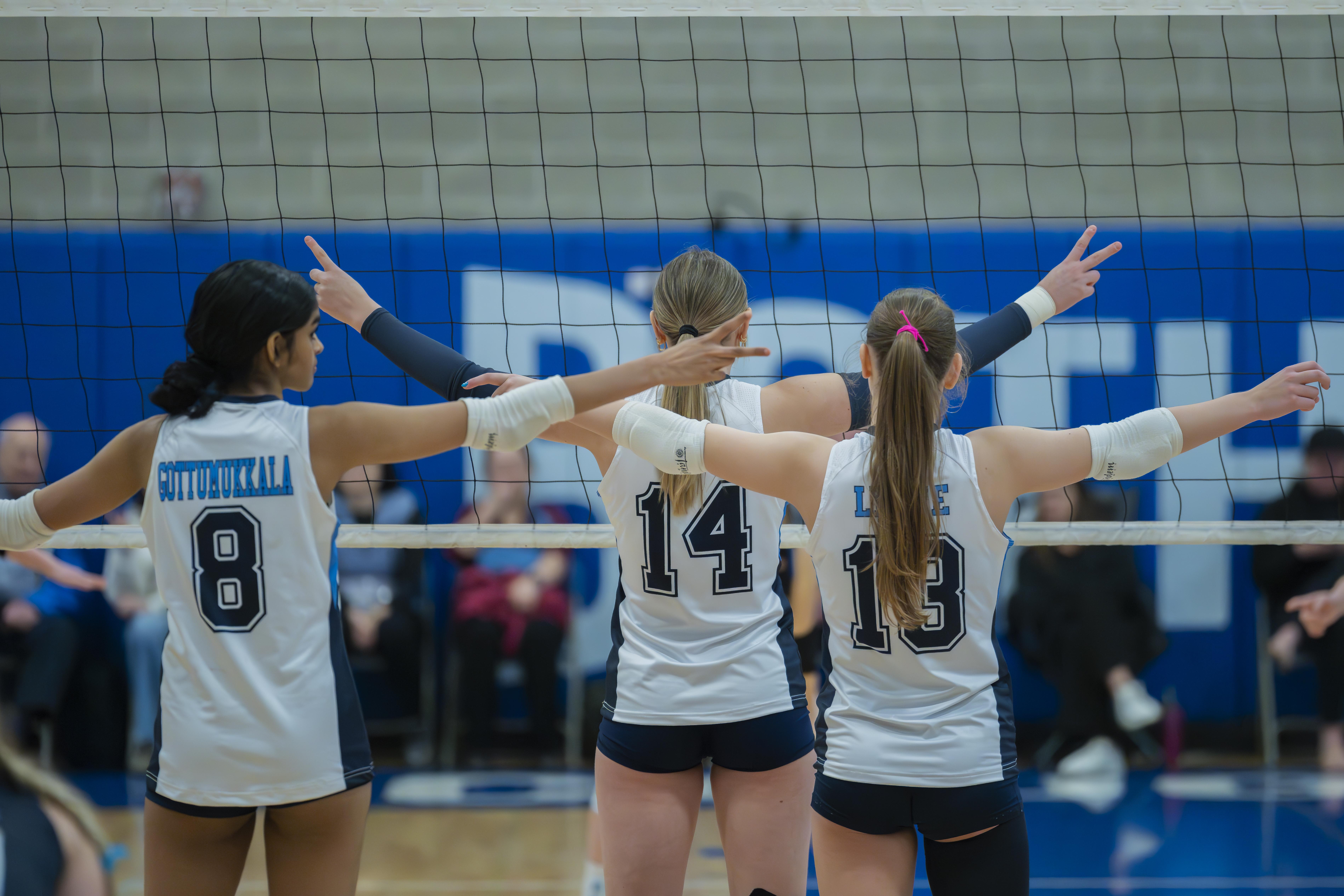 Three volleyball players from the FEVA U15 team playfully pose at the net during a match. Spectators can be seen in the background.