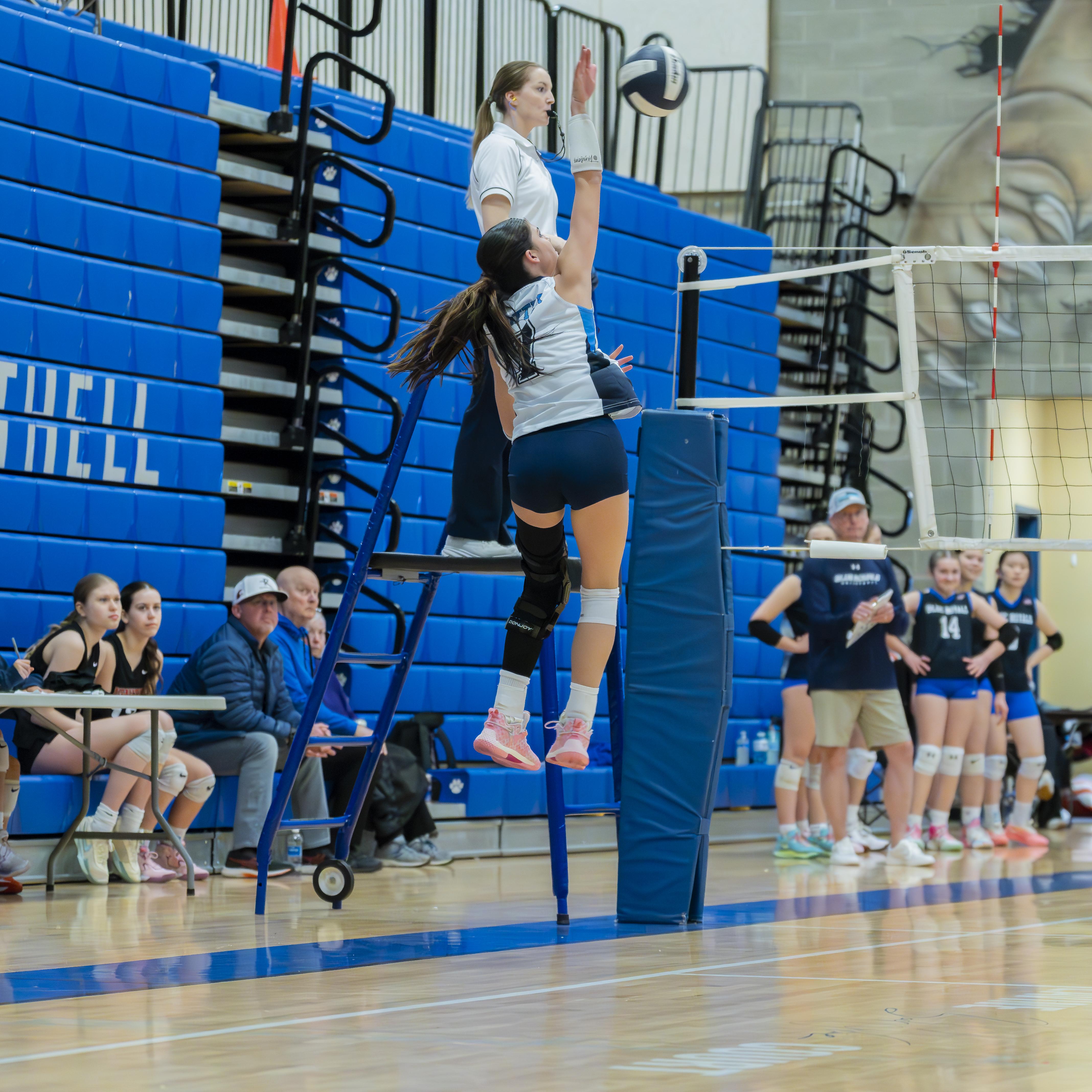 A FEVA volleyball player jumping to spike the ball during a match, while a referee oversees from an elevated position. In the background, teammates and spectators watch attentively.