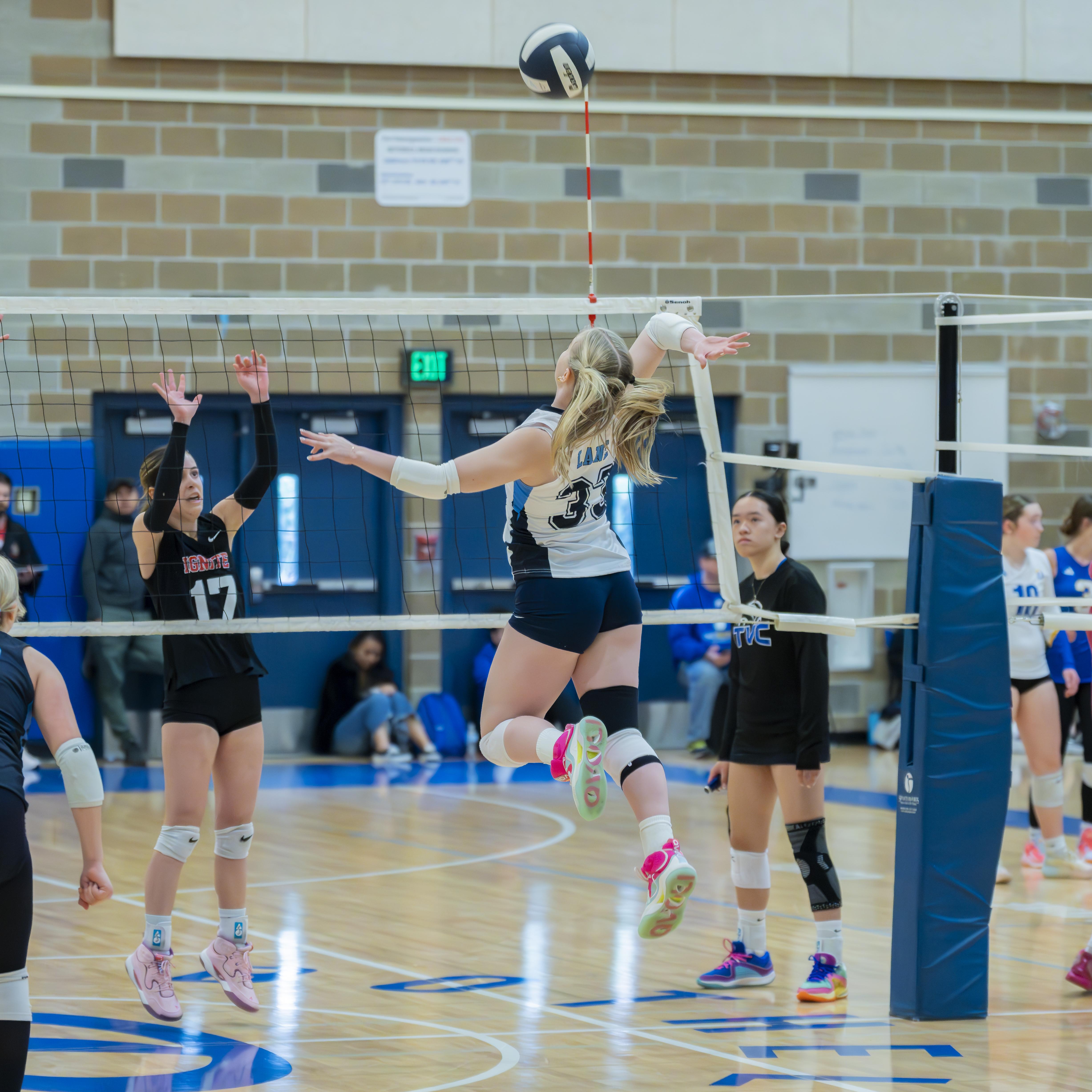 A FEVA volleyball player jumps to spike the ball over the net during a match, while players from the opposing team prepare to block.