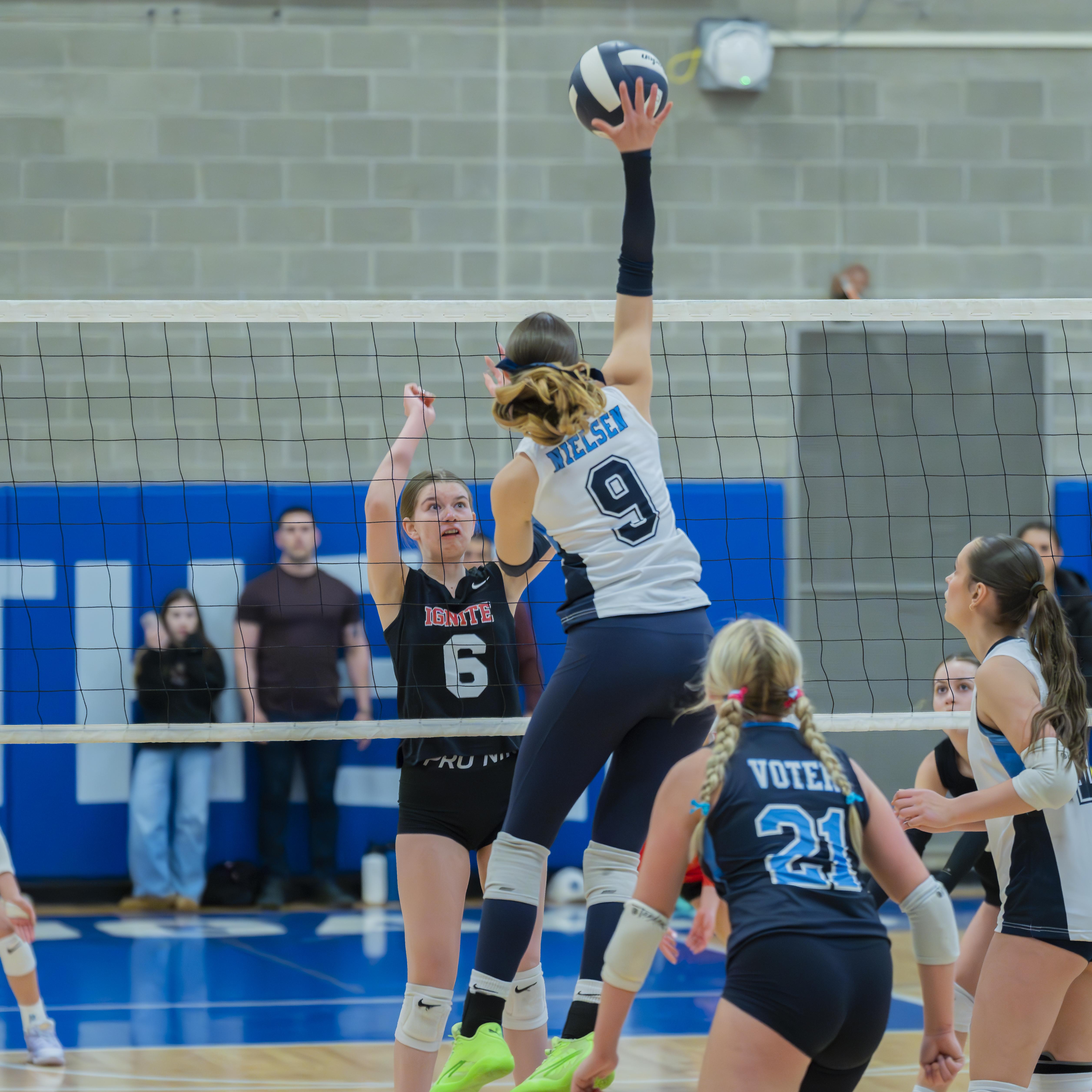 A FEVA volleyball player jumps to set the ball over the net as an opposing player attempts to block, during a match in progress.