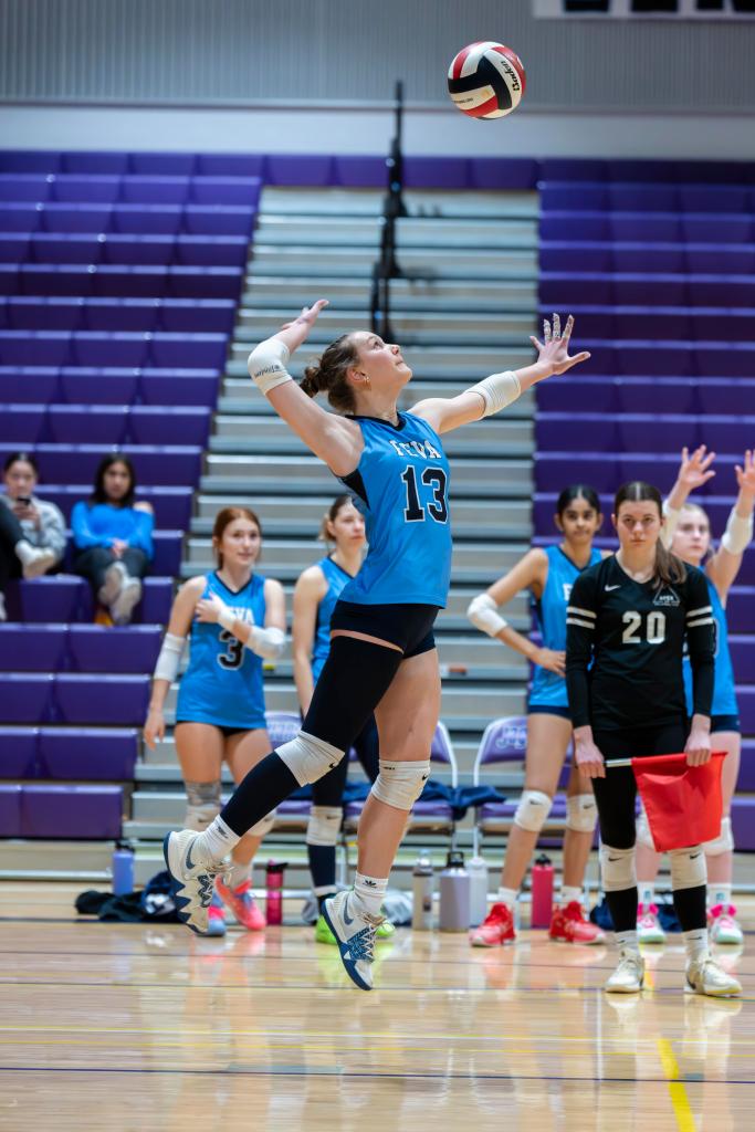A FEVA U15 volleyball player wearing a blue jersey, number 13, jumps to serve the ball during a match, with teammates in the background.