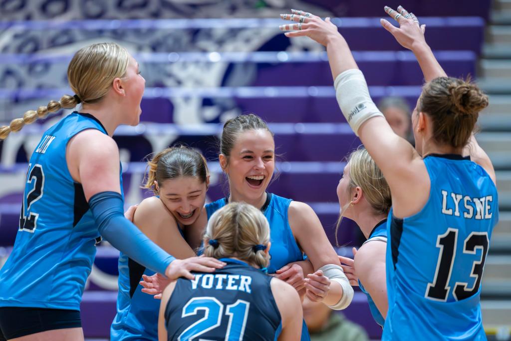 A group of young FEVA female volleyball players celebrating joyfully on the court, wearing blue and black uniforms.