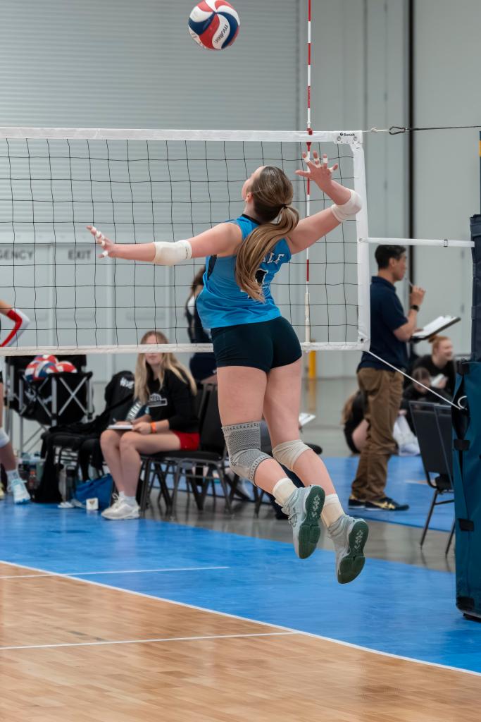 A FEVA U15 female volleyball player leaps to spike a volleyball over the net during a match, wearing a blue jersey and black shorts, with spectators seated in the background.