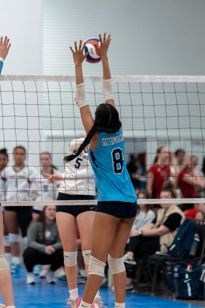 A FEVA U15 female volleyball player in a blue jersey attempts to block a spike at the net during a game, with spectators visible in the background.