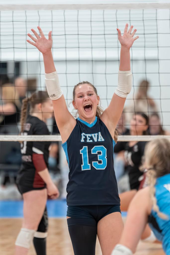 A young FEVA U15 female volleyball player celebrates with her arms raised, wearing a blue jersey with the number 13, during a match. The volleyball net is in the background, and other players are visible in the scene.