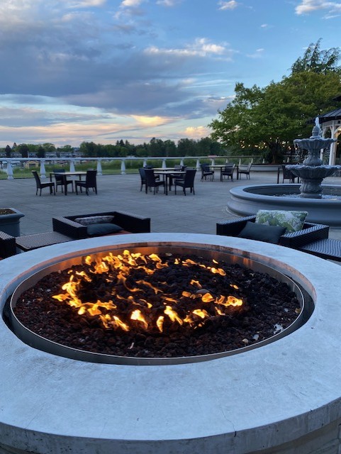 A fire pit with flames surrounded by outdoor seating and tables, set against a background of trees and a beautiful sky.