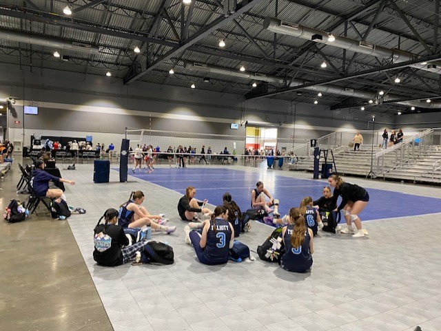 The FEVA U14 volleyball team gathers on the sidelines during a tournament, sitting together in a gymnasium, with players in blue uniforms and a volleyball court in the background.