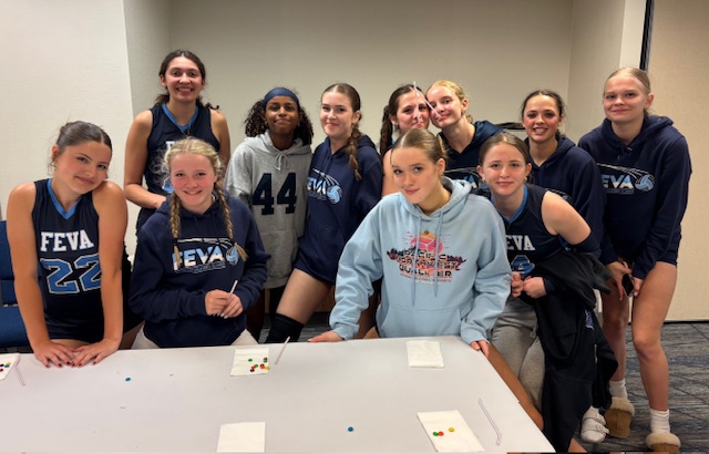 A group of female volleyball players from the FEVA U16 team posing together in a casual setting, wearing team jerseys and hoodies, with a table in front showing snacks.