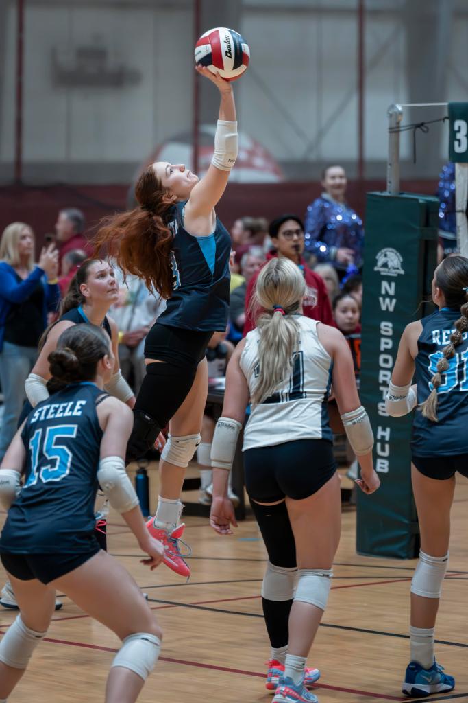 A female FEVA volleyball player jumping to spike a ball during a match, with teammates and spectators in the background.