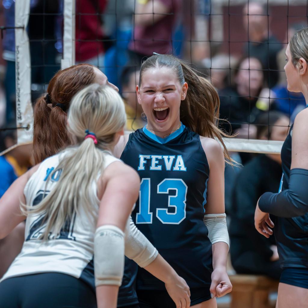 A group of volleyball players celebrating enthusiastically on the court. One player, wearing a black jersey with 'FEVA' and number 13, is joyfully shouting while her teammates express excitement.