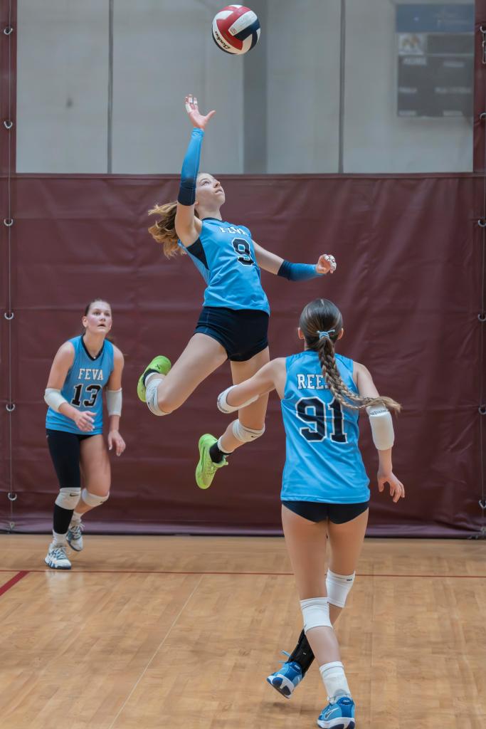A FEVA volleyball player jumps to serve the ball during a game, with two teammates observing in the background.