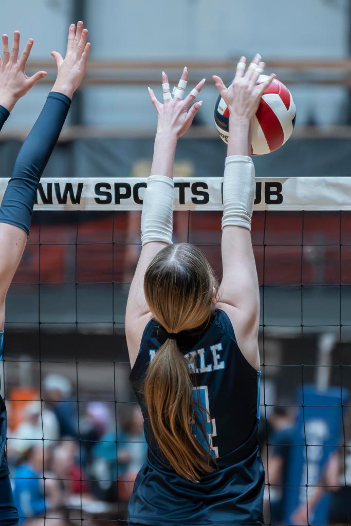 A FEVA volleyball player with long hair jumps to block a ball at a sports club volleyball match, with her hands reaching above the net.