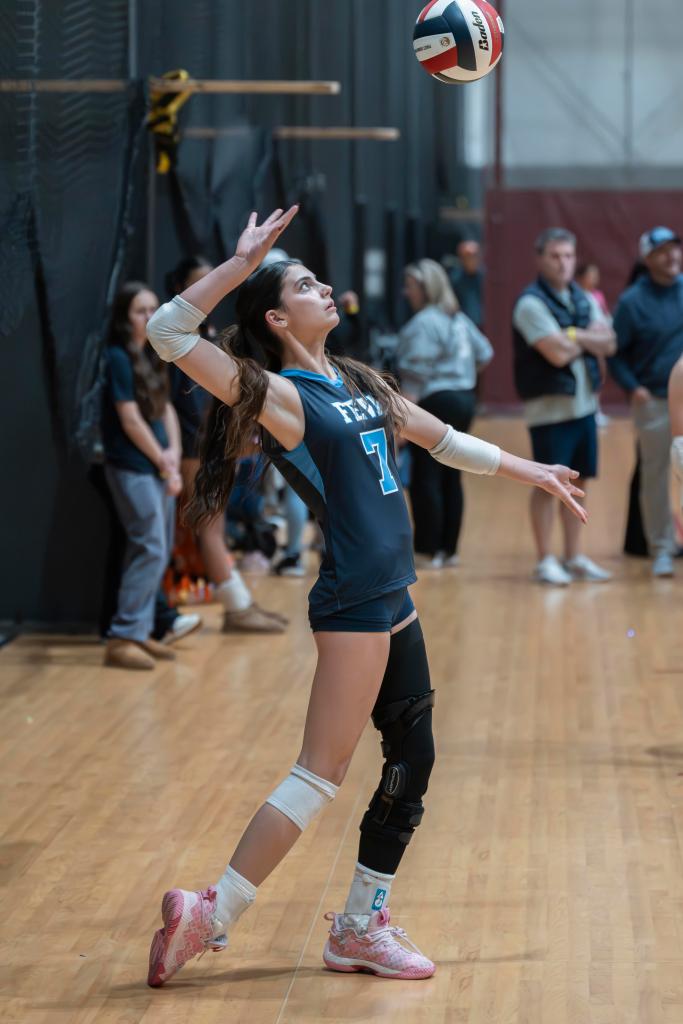 A young FEVA female volleyball player serving the ball during a game. She is wearing a blue uniform with the number 7, bright pink shoes, and a knee brace, with spectators visible in the background.