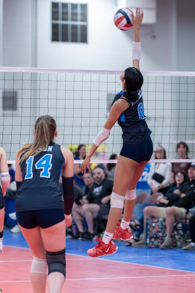 A volleyball player in a blue uniform jumps to spike the ball over the net, while another teammate in the foreground looks on. Spectators can be seen in the background.