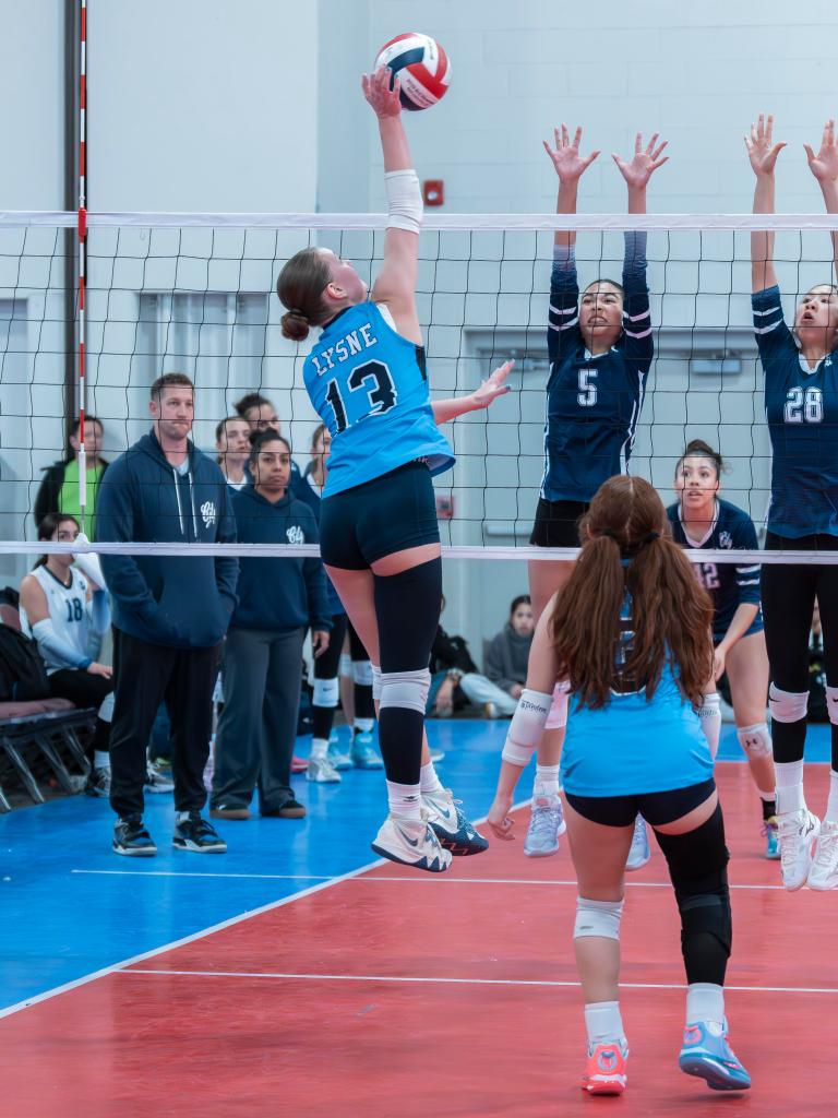 A female volleyball player jumping to spike the ball over the net, with teammates and opponents in the background during a match.