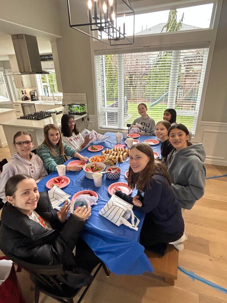 A group of FEVA girls smiling and enjoying a meal together at a table covered with a blue cloth, featuring plates of food and drinks, in a well-lit modern kitchen.