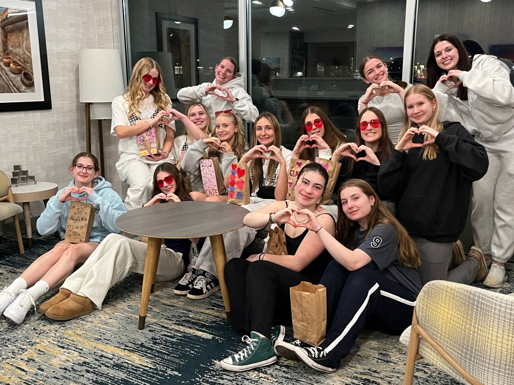 A group of teenage girls in casual clothing posing together in a cozy indoor setting, making heart shapes with their hands, showcasing friendship and camaraderie.