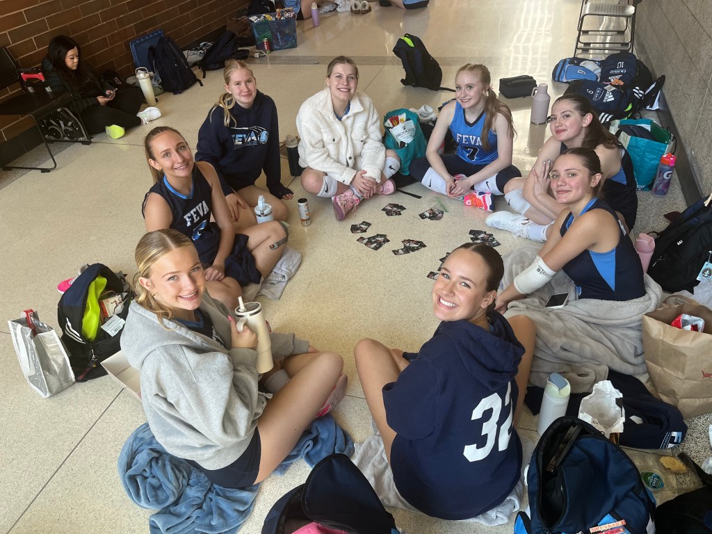 Group of young female volleyball players sitting on the floor, smiling and enjoying time together, surrounded by bags and personal items.