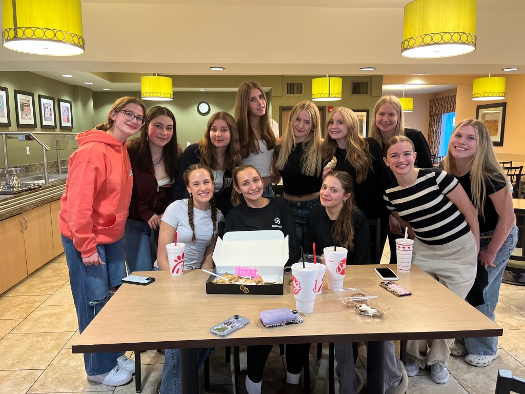 A group of young women posing together at a table in a cozy restaurant, celebrating with food and drinks, showcasing smiles and camaraderie.