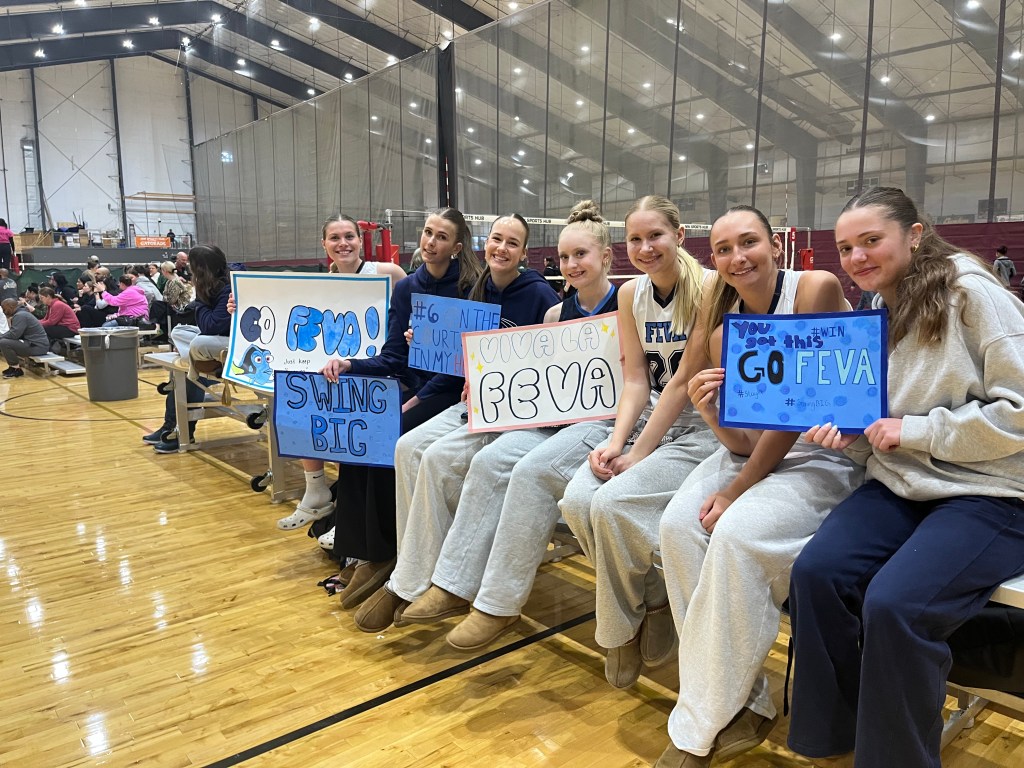 Group of female volleyball players sitting together in a gym, holding supportive signs for their team, FEVA. They are wearing matching sweatpants and jerseys, showcasing camaraderie and team spirit.